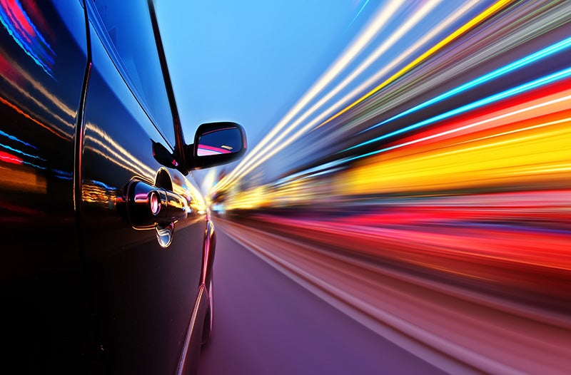 A black car seen from the side, speeding forward with blurred, streaking lights of red, yellow, and blue.